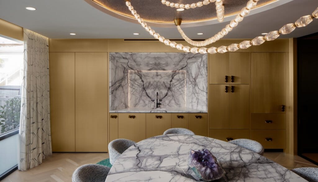 The meeting room at Rob Bowen Design global headquarters with a marble feature wall nested into natural wood cabinetry, a marble conference table, large picture window, textured drapery, and a strung glass chandelier