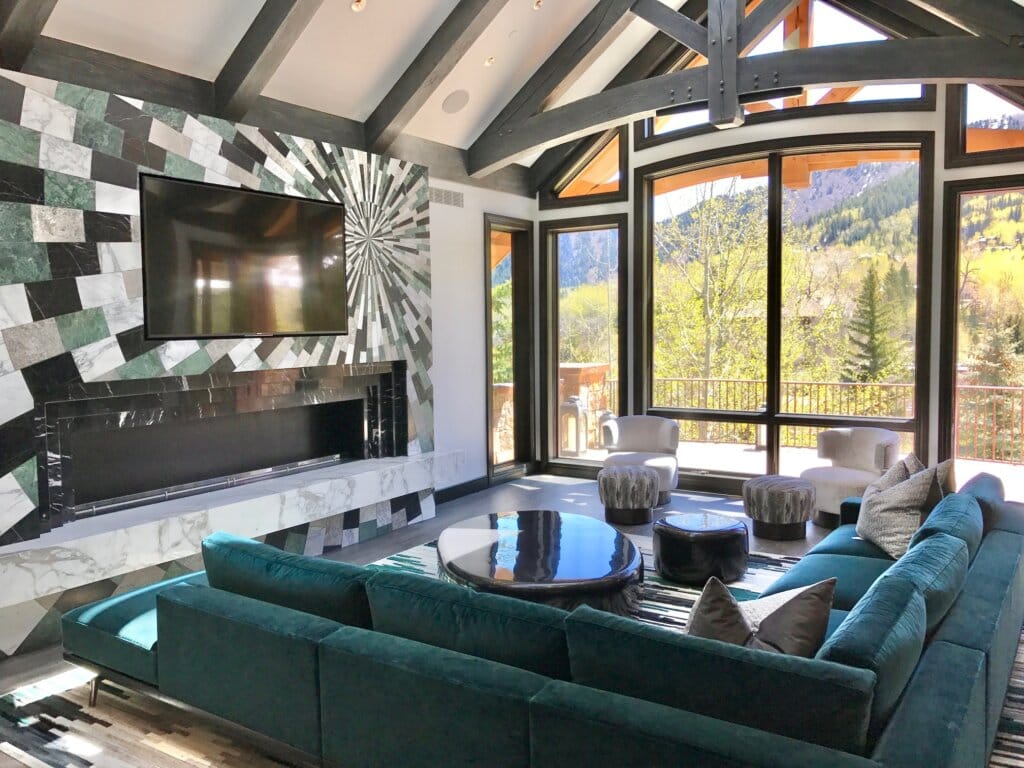 Luxury modern cabin living room design featuring a starburst, checkerboard wall of black, green, grey, and white stone tiles with a flatscreen TV, and a rectangular fireplace. Emerald sectional sofa, black stone oval coffee table, and white accent chairs surround a floor-to-ceiling window facing an expansive forest view. A-frame ceiling with grey-stained beams.