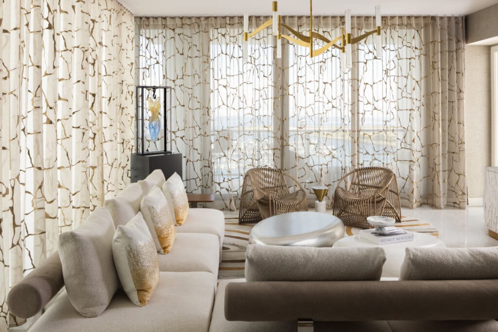 Luxury living room interior design in Tampa Bay featuring sculptural seating and layered textures. Floor to ceiling sheer curtains with gold organic patterns. Modern wicker chairs. 
