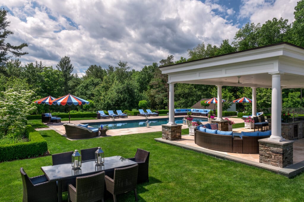 A backyard oasis features a large swimming pool, lounge chairs, umbrellas, and a covered patio with sectional seating and an outdoor dining table.