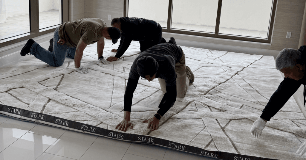 Four individuals are on their hands and knees installing a large, light-colored rug with a geometric pattern into a South Florida high-rise luxury condo. The rug has a dark border with the word "STARK" repeated multiple times. 