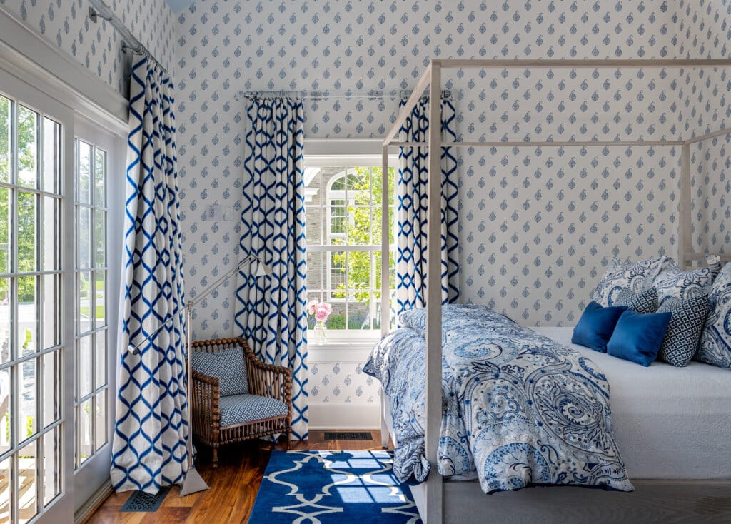 A luxury pool house bedroom with blue and white patterned wallpaper and curtains, a four-poster bed with matching bedding, and a wicker chair.