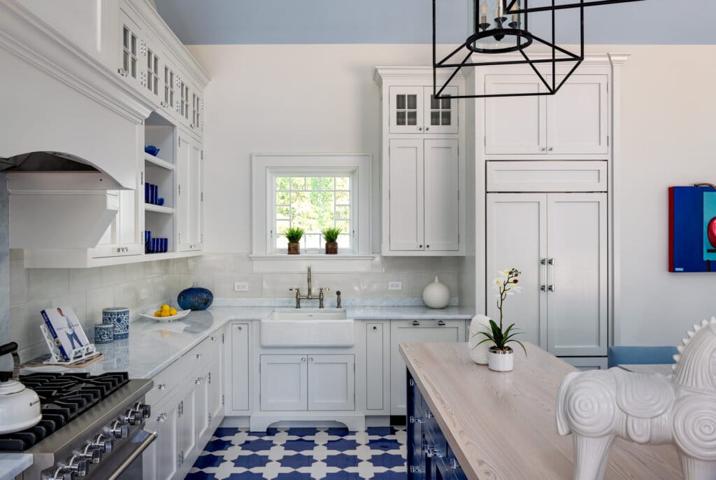 A luxury pool house bright white kitchen with blue and white tile flooring, white cabinets, a farmhouse sink, and a large island with a wooden countertop. A modern black chandelier hangs from the blue ceiling.