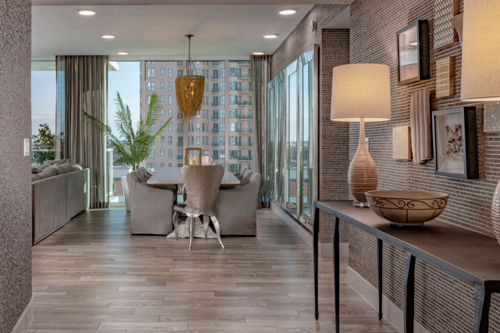 A dining room and living area with large windows overlooking a cityscape. Wooden table with upholstered chairs, and a woven chandelier. A console table with decorative lamps and a bowl is against a textured wall.