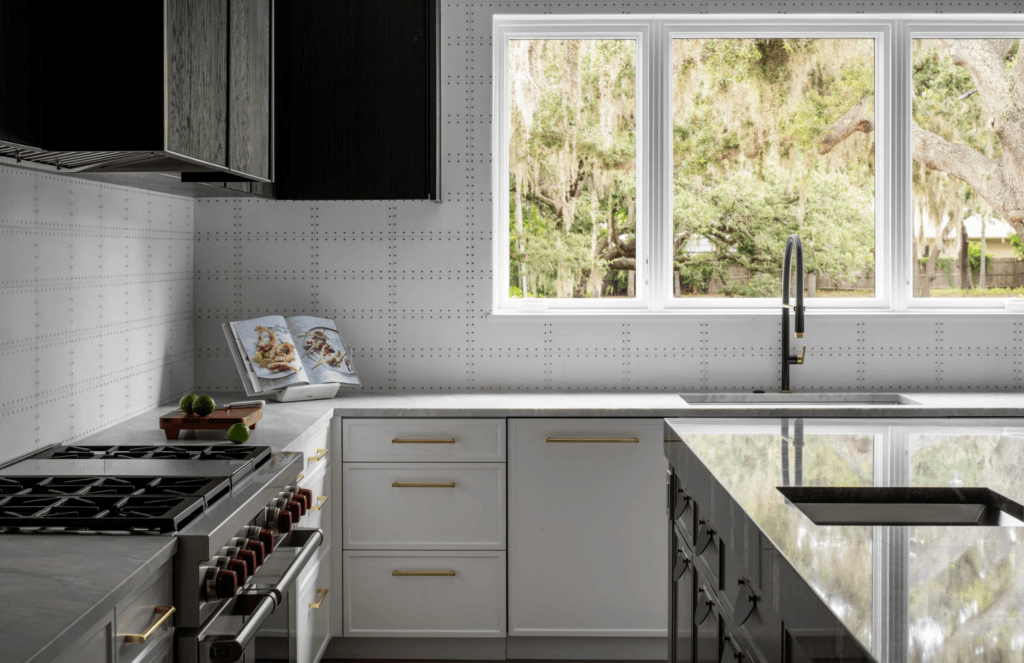 A modern kitchen with a stainless steel range, white cabinets with gold hardware, and a large window overlooking trees with Spanish moss.