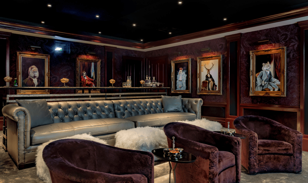 A luxurious home theater room features a tufted leather sofa, plush purple armchairs, and ornate framed portraits of animals in formal attire on a damask-patterned wall.