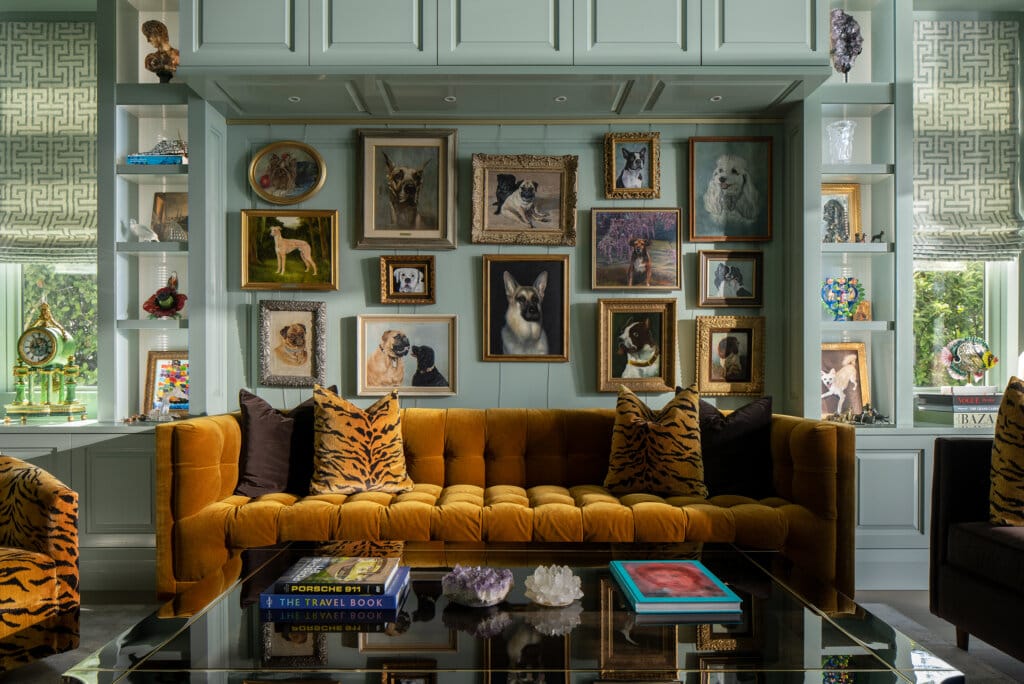 A tufted gold sofa with tiger-print pillows is in front of a wall adorned with numerous framed dog portraits. A glass coffee table holds books and crystals.