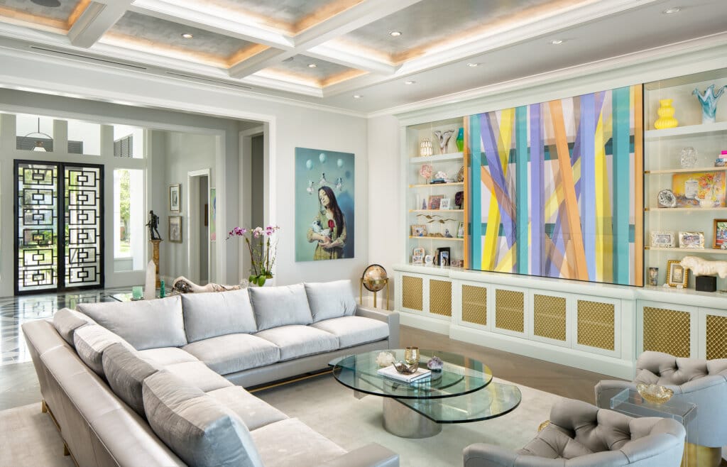 A bright and airy living room with a large sectional sofa, glass coffee table, and a built-in wall unit displaying art and decorative objects and abstract paintings on the wall.
