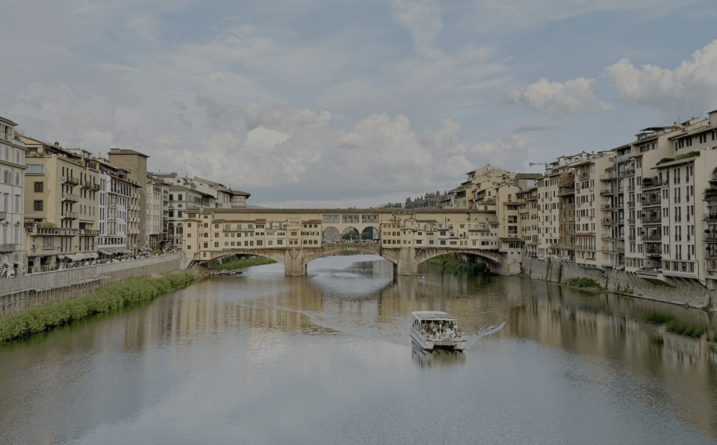 A boat carrying passengers travels down the Arno river lined with old buildings, with the Ponte Vecchio  in the background under a blue sky with white clouds.