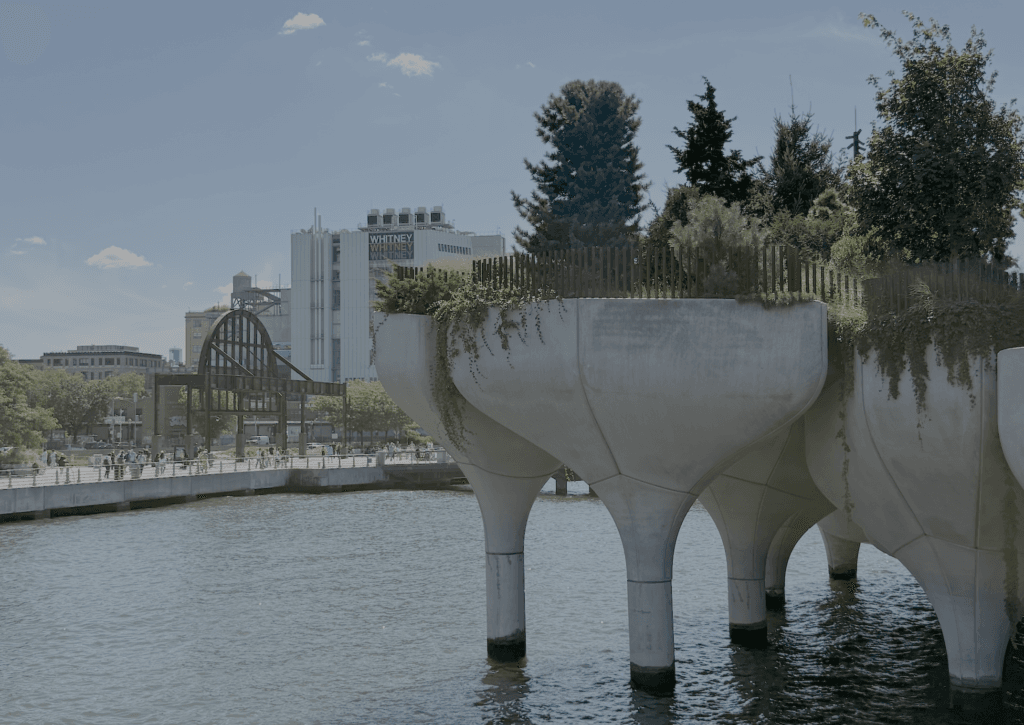 Little Island, public park in New York City. Tulip-shaped concrete structures over the Hudson river with trees, with buildings and a walkway visible in the background.