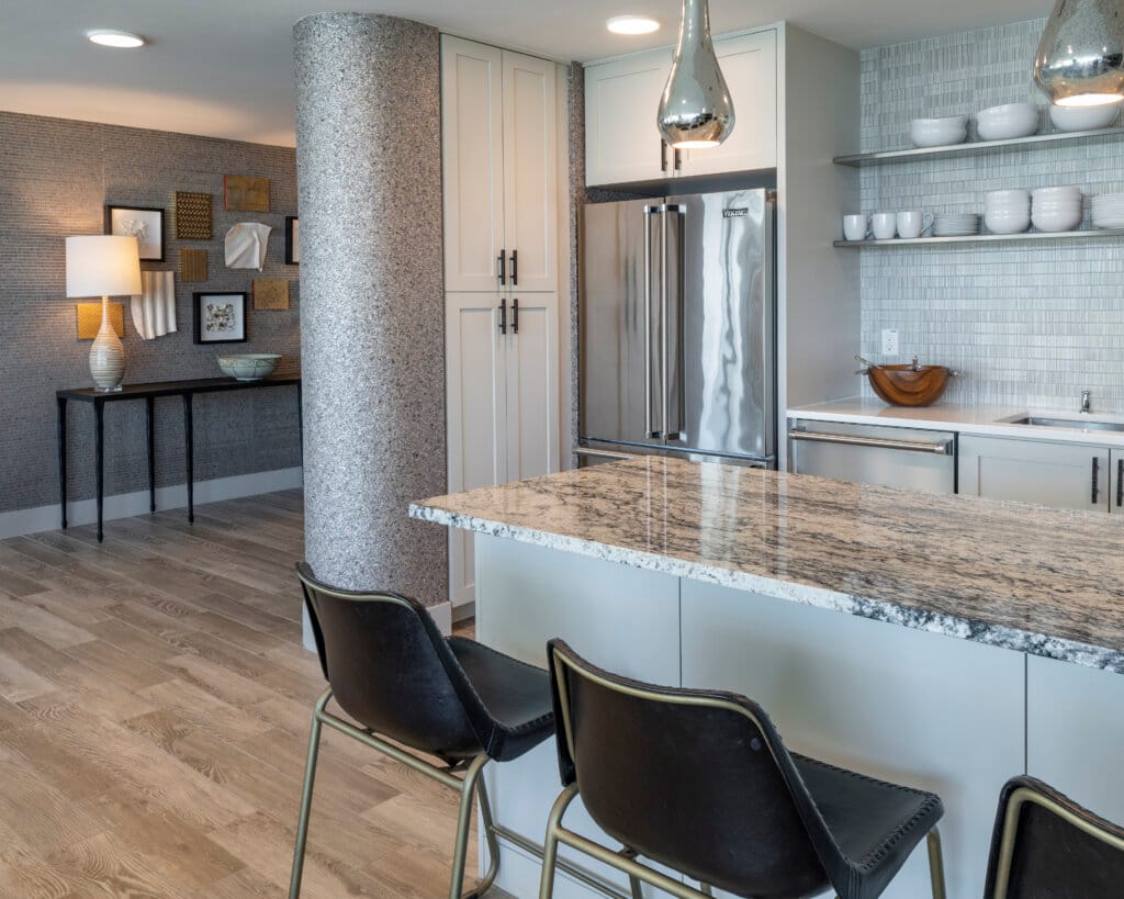 A modern kitchen features a granite-topped island with bar stools, a stainless steel refrigerator, and a tiled backsplash with open shelving.