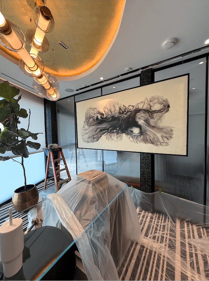 A painting of abstract ink swirls hangs on a wall above a piece of furniture covered in plastic sheeting, with a ladder nearby and a large potted plant to the left.