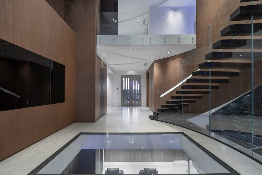 A modern hallway with a floating staircase, glass railings, and a glass floor section revealing a lower level with seating.