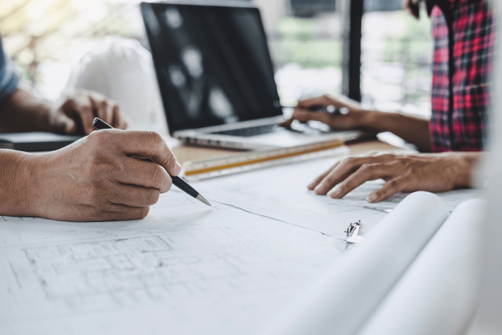 Two people reviewing blueprints at a table with a laptop and ruler.