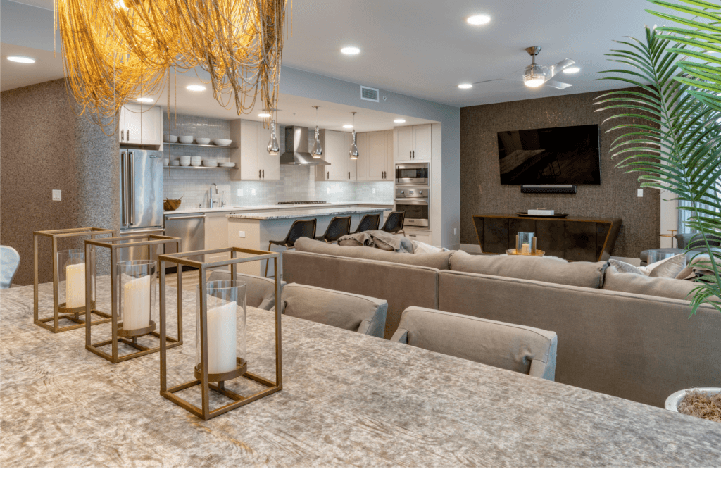 A spacious open-plan living and kitchen area with a large sectional sofa, a sleek modern kitchen with stainless steel appliances, and a striking gold chandelier above a dining table with decorative candles.