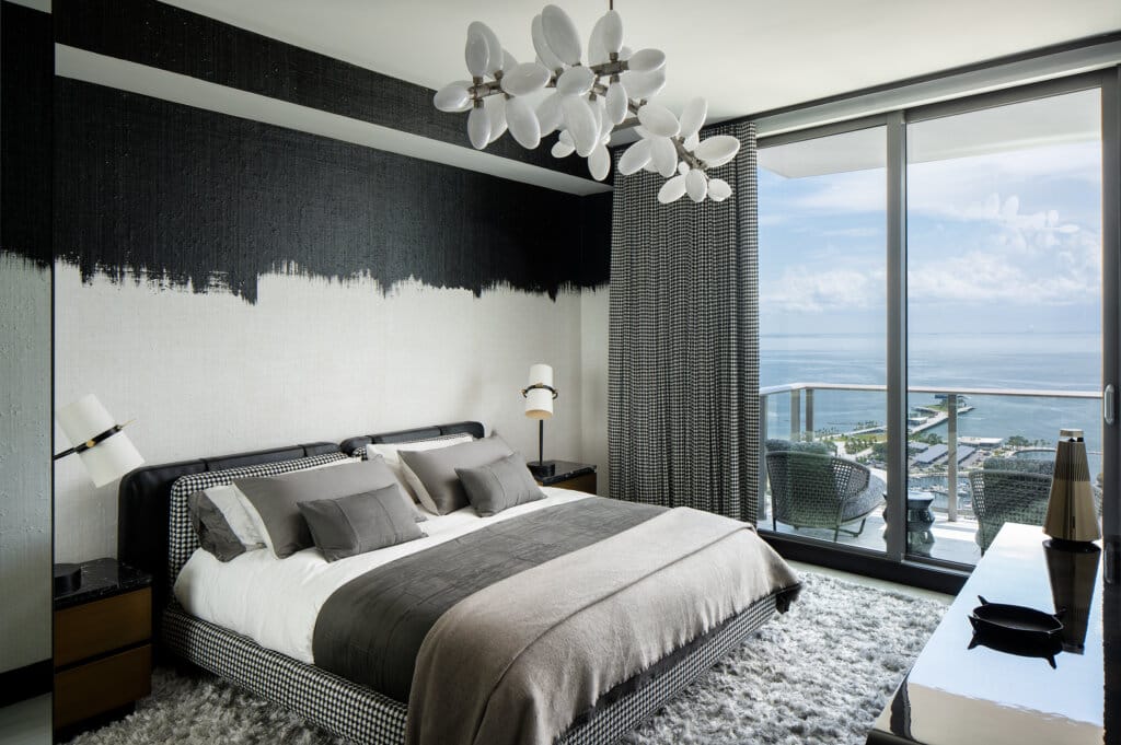Modern bedroom with a large bed, dark headboard wall, patterned curtains, a grey rug, and a chandelier above; floor-to-ceiling windows show an ocean view and balcony railing.