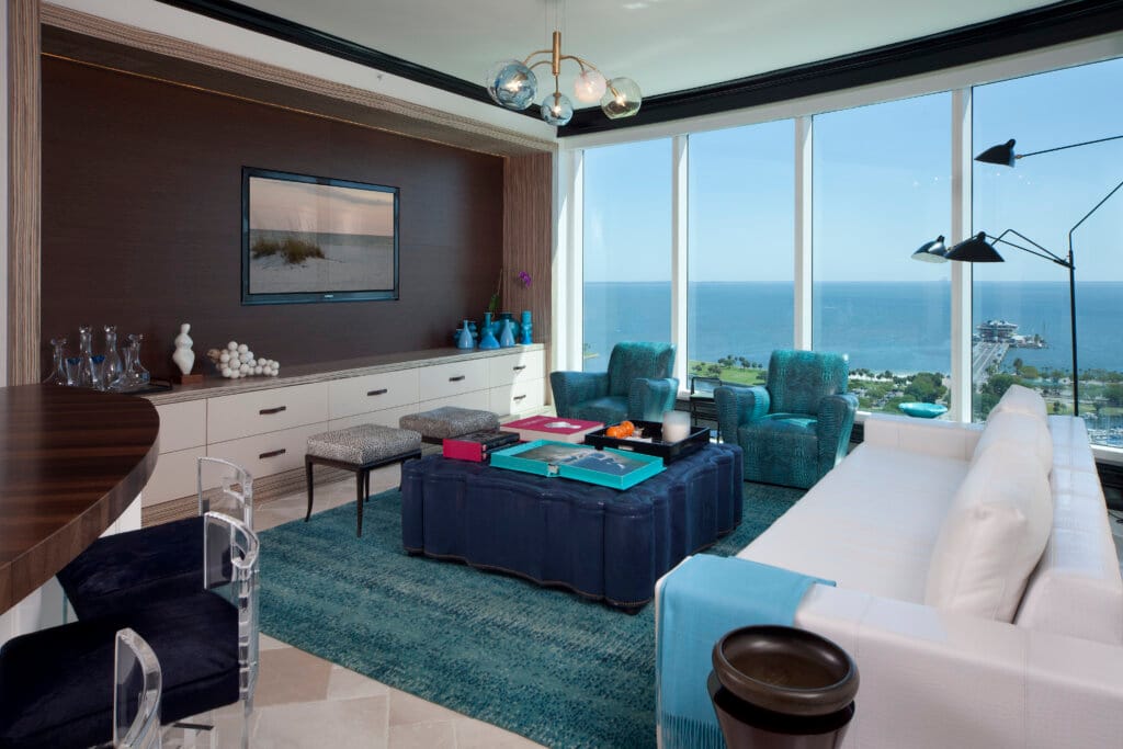Upscale living room with teal armchairs and a dark coffee table, large windows overlooking the ocean, a chandelier overhead, framed wall art, and a bar counter on the left.