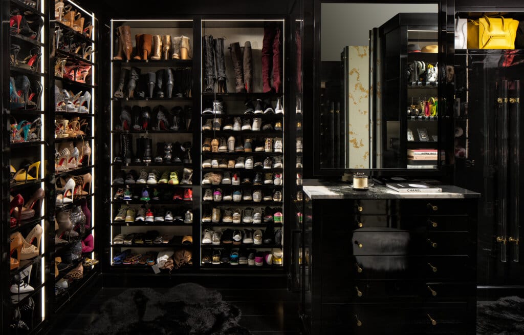Luxury interior with multiple built-in glass cabinets displaying rows of shoes and accessories, plus a dark vanity and mirror with gold hardware in a dim, moody lighting setup.