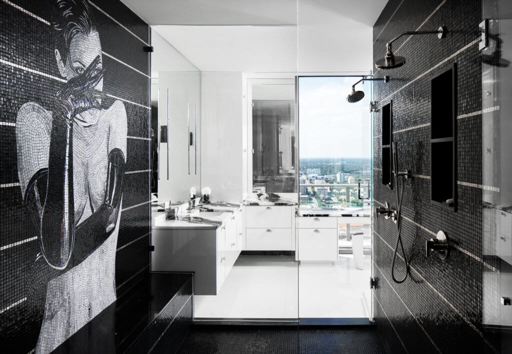 Modern black-and-white bathroom with dark tiled walls, two sinks and cabinets, a shower area with glass doors, and a ceiling light fixture, leading to a bright hallway with a city view through the window.