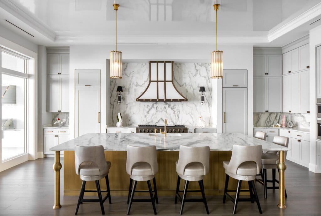 Bright modern kitchen with a large marble island and five bar stools, white cabinetry, marble backsplash with fireplace feature, and three gold pendant lights hanging over the island.